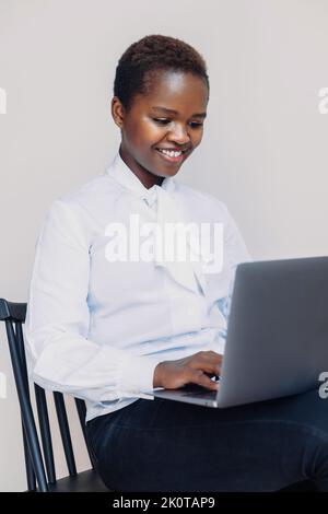 Femme afro-américaine portant une chemise dactylographiée à l'aide d'un ordinateur portable pour naviguer sur Internet assis sur une chaise contre un mur blanc. Fille lisant blog Banque D'Images