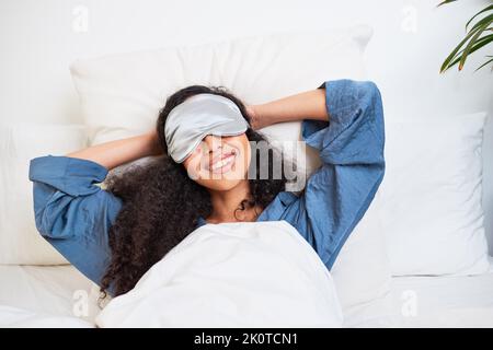 Une jeune femme multiethnique s'étire avec les bras derrière la tête et le masque de sommeil Banque D'Images