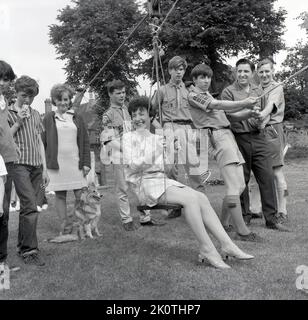 1967, historique, à l'extérieur dans un champ à une fête de village, une excité - ou perhas nerveux - dame en jupe courte de l'époque, sur le point d'avoir un tour sur un télésiège de corde étant géré par des scouts, Stone, Buckinghamshire, Angleterre, Royaume-Uni, regardé par des jeunes locaux. Banque D'Images