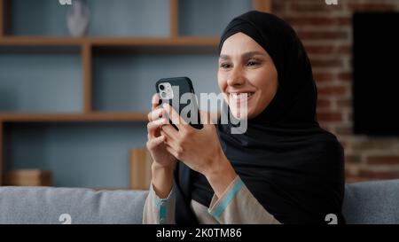 Femme musulmane détendue dans le hijab noir utilisant un smartphone à la maison avec le sourire surfant sur les médias sociaux dans l'application mobile de vérification des nouvelles d'achat de nourriture Banque D'Images