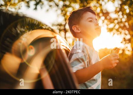 Un petit garçon gai en train de profiter de la promenade avec ses parents en voiture en été, tout en mangeant un en-cas. Voyage en famille le week-end, vacances ensemble, route et Banque D'Images