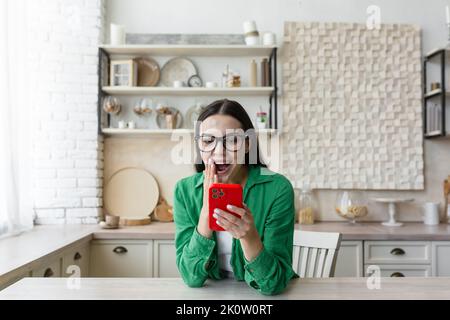 Jeune femme satisfaite belle en lunettes et une chemise verte à la maison dans la cuisine tenant un téléphone rouge dans ses mains, heureux acheteur en ligne, shopping réussi. Banque D'Images