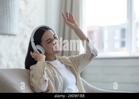 Belle jeune femme écoutant de la musique dans un casque blanc. Il est assis à la maison sur le canapé avec ses yeux fermés, danse, chante, fait des vagues. Banque D'Images