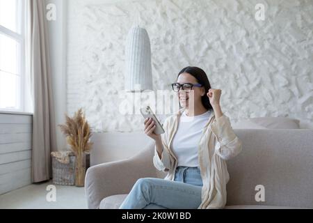 Bonne jeune femme assise sur un canapé à la maison et tenant le téléphone. Bonne nouvelle. A obtenu un emploi réussi, a passé un entretien, a reçu un message. Il se réjouit, crie avec joie, pointe avec sa main oui. Banque D'Images