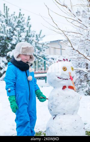 Enfant, garçon debout bonhomme de neige et le tenant avec la main. Bonhomme de neige construit dans l'arrière-cour couvert de neige. Bonne enfance. Hiver à la campagne. Banque D'Images