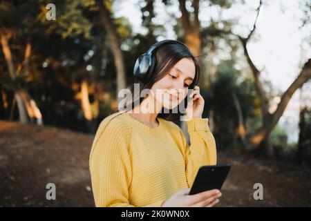 Adolescente portant un chandail jaune, utilisant un casque pour jouer de la musique sur son smartphone dans le parc Banque D'Images