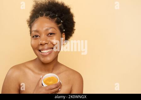 Charmante gaie mauriquement africaine femme américaine tient des taches cosmétiques sous ses yeux, regarde joyeusement la caméra, pose sur un fond beige, copie Banque D'Images