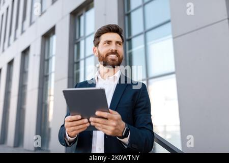 Heureux confiant, beau jeune homme caucasien avec barbe en costume dactylographiant sur la tablette, regardez l'espace vide Banque D'Images