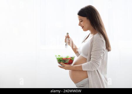 Femme enceinte affamée qui mange de la salade de légumes saine, vue latérale Banque D'Images
