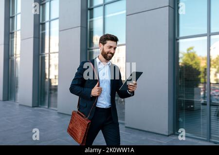 Joyeux jeune homme caucasien confiant avec la barbe en costume bavardant sur tablette, va au travail Banque D'Images