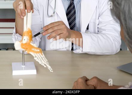 Chirurgien orthopédique en robe blanche et stéthoscope pointant vers le squelette humain pied cheville os modèle anatomique d'articulation, présente au patient âgé environ Banque D'Images