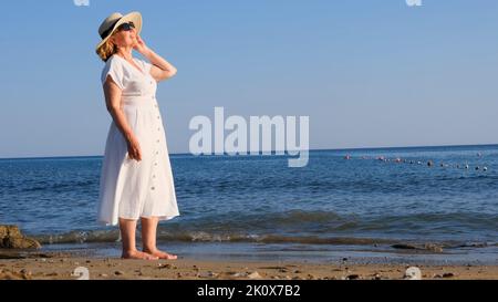 femme en chapeau de paille et robe blanche marche le long de la mer et admire la mer bleue lors d'une journée ensoleillée d'été, profitant de la liberté et de la détente.Le Banque D'Images