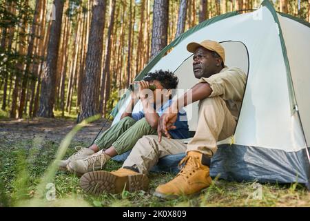 Adorable garçon afro-américain regardant à travers des jumelles tout en étant assis dans une tente à côté de son grand-père en vêtements décontractés pendant la randonnée dans la forêt Banque D'Images