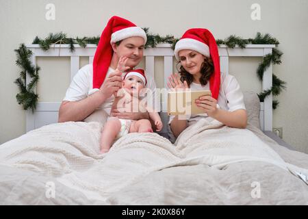 Mari et femme avec bébé communiquent par tablette sur le lit décoré pour noël et nouvel an, couple de famille dans la chambre à la maison - Moscou, Russie, Decembe Banque D'Images