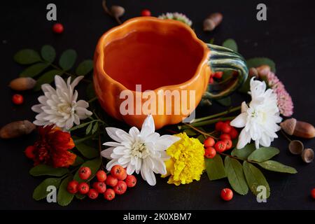 Tasse de thé d'automne en forme de citrouille parmi les fleurs : fond avec baies de rowan, glands, marigold, chrysanthème sur fond noir Banque D'Images