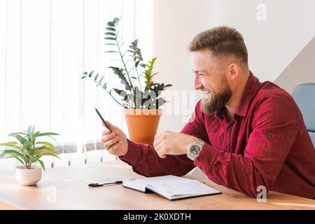 Un journaliste indépendant rouge et décontracté qui a réussi à discuter de la communication vidéo par téléphone tout en étant assis à son bureau et en prenant des notes en riant avec rire. Vidéoconférence avec les partenaires professionnels Banque D'Images