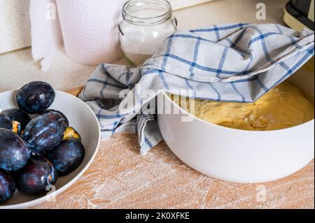 Prune sur assiette, pâte de levure dans un bol avec serviette, verre avec sucre et essuie-tout de cuisine en papier sur planche de bois arrosée de farine, gros plan. Préparation du sucré Banque D'Images