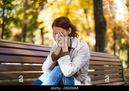 Une femme blanche en imperméable aux cheveux rouges, desappariée, est assise sur le banc et ferme les yeux avec les mains, pleure dans le parc Banque D'Images