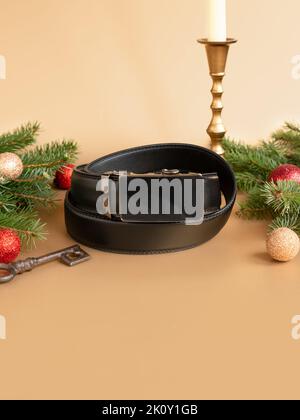 Ceinture en cuir roulée noir tendance pour hommes avec boucle automatique, sapin de Noël, boules brillantes, bougeoirs dorés et clé sur fond doré. Banque D'Images