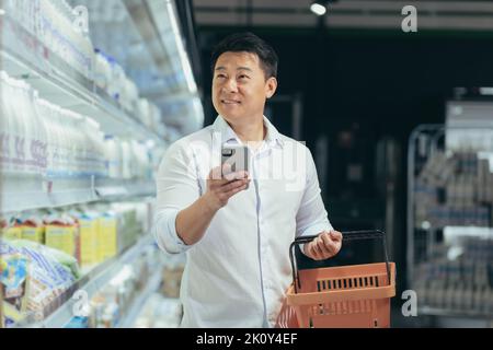 un acheteur d'homme asiatique se promène dans le supermarché avec un panier d'achat regardant le smartphone en naviguant dans les mains rechercher des biens ou des produits sur la liste client acheteur mâle dans une épicerie. Banque D'Images