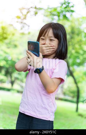 Belle fille asiatique utilisant un smartphone mobile. Petite fille joue dans le jeu numérique dans le parc vert. Banque D'Images