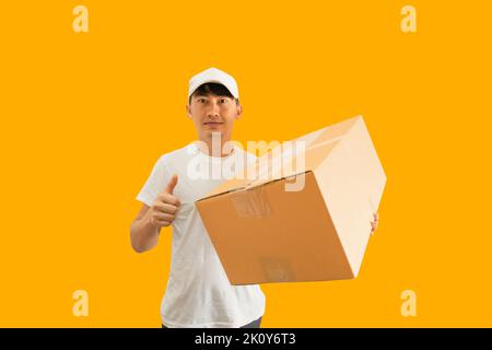 Jeune homme de livraison asiatique portant une casquette et un t-shirt blanc vierge tenant une boîte postale isolée sur fond jaune. concept de service de livraison express. Banque D'Images