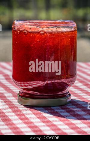 Un pot en verre avec confiture de fraises maison fraîche debout à l'envers sur une table avec une nappe à carreaux rouges. Banque D'Images