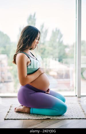 Portrait de la jeune femme enceinte dans des vêtements de sport assis contre la grande fenêtre tenant les mains sur son ventre. Grossesse, attente et tendresse concept. C Banque D'Images