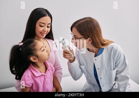 Docteur ophtalmologiste utilisant l'ophtalmoscope regardant dans les yeux de la petite fille asiatique qui est assise à côté de sa mère. Contrôler la vision de l'enfant Banque D'Images