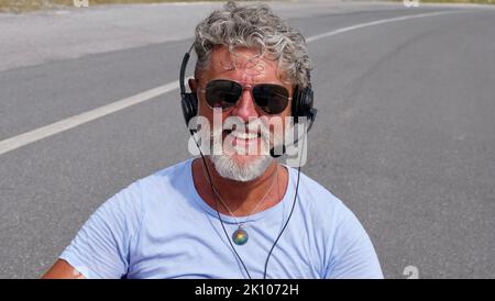 Homme âgé aux cheveux gris indépendant avec une barbe en lunettes de soleil, travaillant dans les écouteurs sur la route. Un vieil homme fou et extraordinaire en été Banque D'Images