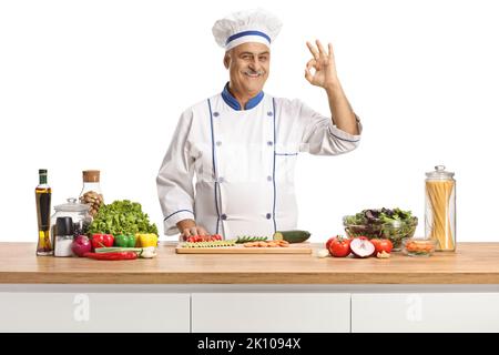 Chef mâle mature posant derrière un comptoir avec des légumes et gesturant bon isolé sur fond blanc Banque D'Images