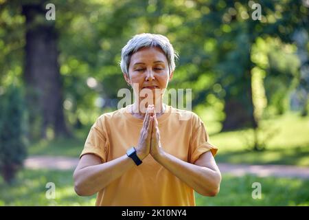 Femme asiatique active plus ancienne pratiquer la méditation avec les yeux fermés poser dans le parc d'été bras pliés fait mudra ou signe Namaste, faire l'entraînement de yoga dehors Banque D'Images