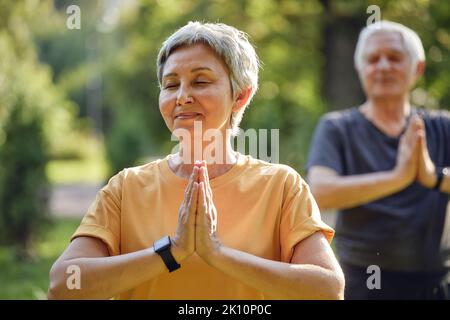 Couple actif plus âgé pratiquer la méditation avec les yeux fermés poser dans le parc d'été bras pliés fait mudra ou signe Namaste, faire l'exercice de yoga dehors, lui Banque D'Images
