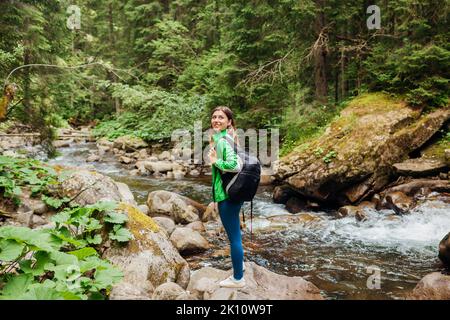 Voyageur randonneur avec sac à dos apprécie le paysage de la rivière de montagne dans la forêt de Carpathian. Une jeune femme marche sur des rochers. Voyage à l'été Ukraine Banque D'Images
