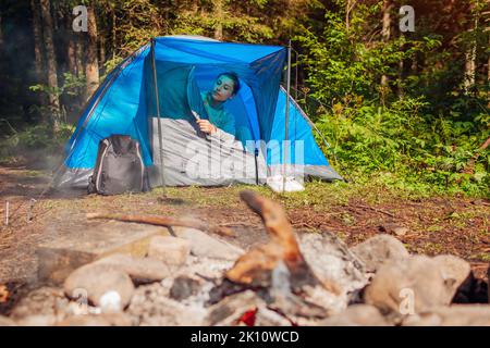 Jeune femme se réveillant la tente de dézipping par un feu de camp dans la forêt d'été le matin. Voyageur en voyage de loisirs. Camping Banque D'Images