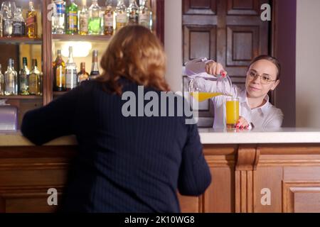 Jeune serveuse en verres versant du jus dans le verre de la carafe à une femme méconnaissable, assis au bar du restaurant Banque D'Images