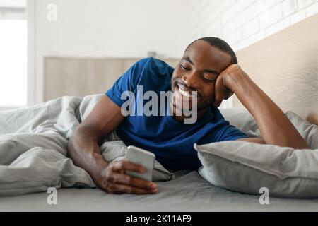 Happy Black Guy utilisant le téléphone dans le lit SMS dans la chambre Banque D'Images