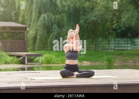 Une belle femme assise sur une plate-forme en bois près d'un étang sur tapis en été, fait du yoga Banque D'Images
