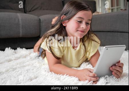 Adorable petite fille montre des dessins animés sur une tablette numérique. L'enfant se trouve sur le sol en riant à l'aide d'un appareil électronique. Loisirs en intérieur pour les enfants. Photographie de haute qualité. Banque D'Images