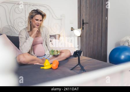 Femme enceinte du caucase moderne assise sur un lit, mangeant une salade nutritive et grignotant des fruits, parlant à ses amis lors d'un appel vidéo. Photo de haute qualité Banque D'Images