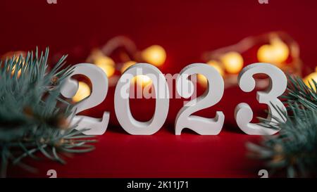 Bonne année 2023. Support en bois blanc numéros 2023 avec branches d'arbre de Noël sur fond rouge avec ampoules guirlande. Joyeux Noël. Banque D'Images