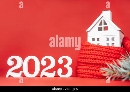 Bonne année 2023. Support en bois blanc numéros 2023 avec maison blanche de jouet enveloppée dans une écharpe rouge et branche d'arbre de Noël sur fond rouge. Joyeux Banque D'Images