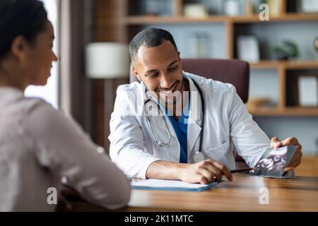 Souriant, un médecin gynécologue afro-américain montrant une image échographique de la patiente Banque D'Images