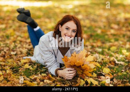 Bonne jolie souriante caucasienne millénaire rouge-cheveux dame en imperméable avec des feuilles jaunes dans les mains se trouve sur le sol Banque D'Images