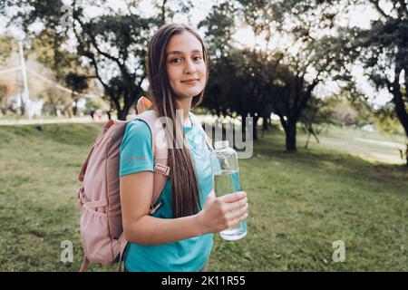 Adolescente portant une chemise turquoise et un sac à dos rose, tenant une bouteille de verre avec de l'eau dans la nature Banque D'Images