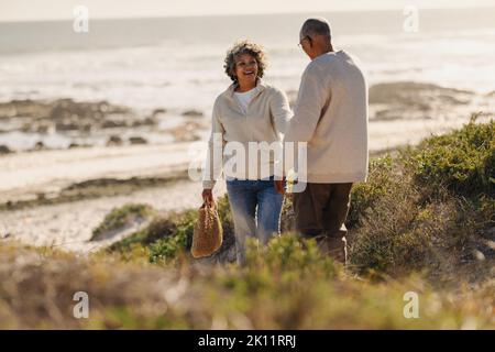Bonne femme âgée tirant son mari par la main tout en allant à la plage pour un pique-nique. Couple senior joyeux prenant des vacances en bord de mer après avoir pris sa retraite Banque D'Images