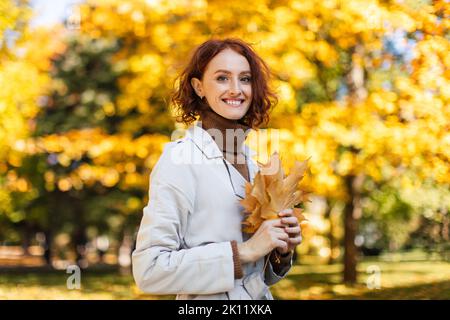 Marche et mode de vie actif. Bonne femme caucasienne à tête rouge millénaire en imperméable tient des feuilles jaunes Banque D'Images