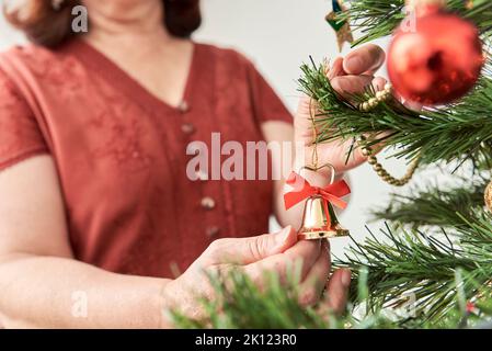 Femme méconnaissable décorant un arbre de Noël à la maison, accrochant une cloche sur une branche. Composition lumineuse avec espace de copie. Banque D'Images