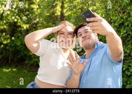 Un jeune couple asiatique de chubby prend ensemble une photo de selfie dans le parc vert à l'extérieur. Un homme et une femme souriants qui appellent quelqu'un sur un smartphone Banque D'Images