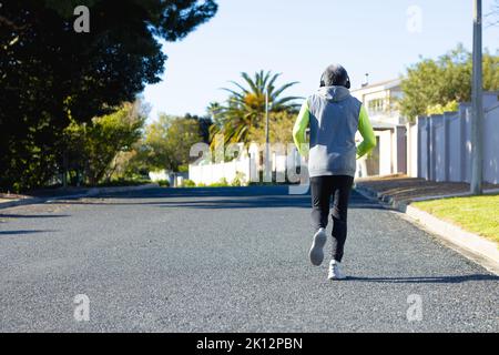 Vue arrière de l'homme biracial senior en vêtements de sport et casque de jogging sur route ensoleillée Banque D'Images